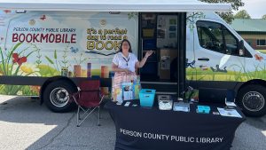 Dawn Gaug, a Person County Public Library staff member, stands beside a book display and outreach table in front of the colorful library bookmobile.