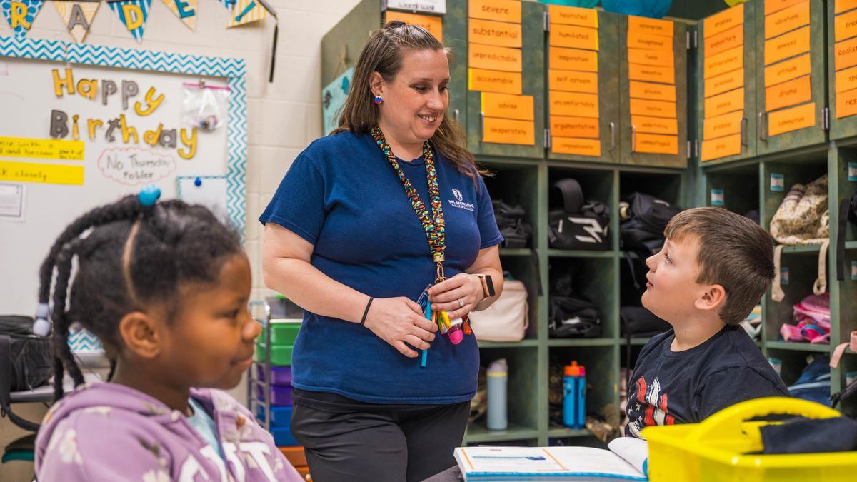 An elementary school teacher speaks with a student at a classroom table while another student sits nearby.