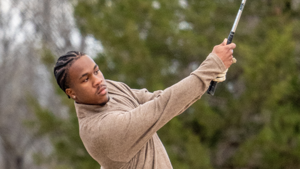 Cameron Davis follows through on a golf swing outdoors, wearing a tan long-sleeve shirt with trees blurred in the background.