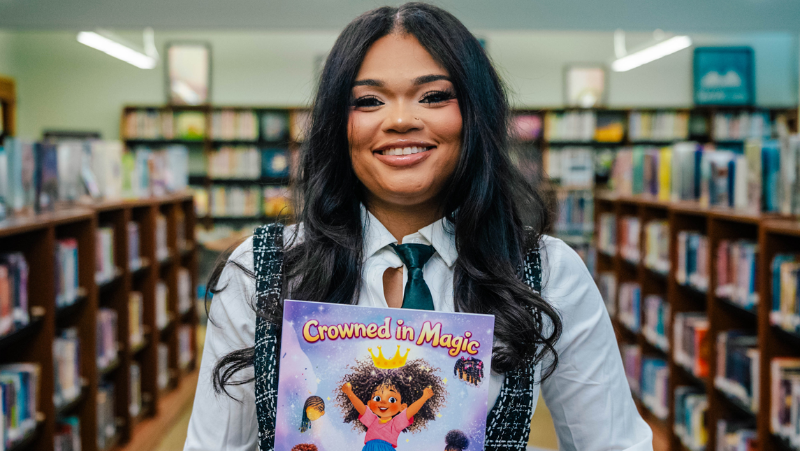 Naiyana Thues smiles in a library while holding a copy of Crowned in Magic.